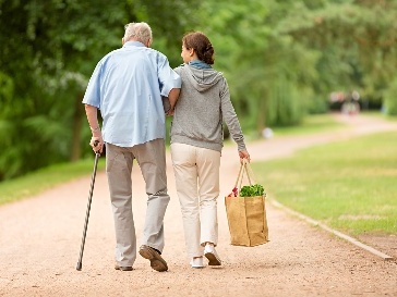 2 people walking in a park. One is older and using a walking stick. The other is supporting them and carrying a bag.